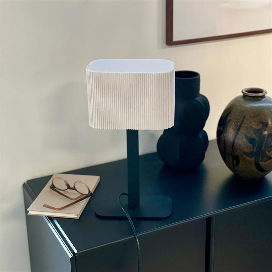 Table lamp with white, grooved lampshade stands on a dark sideboard; wire runs down, a pair of glasses rests on a notebook to the left, two ceramic vases stand behind.