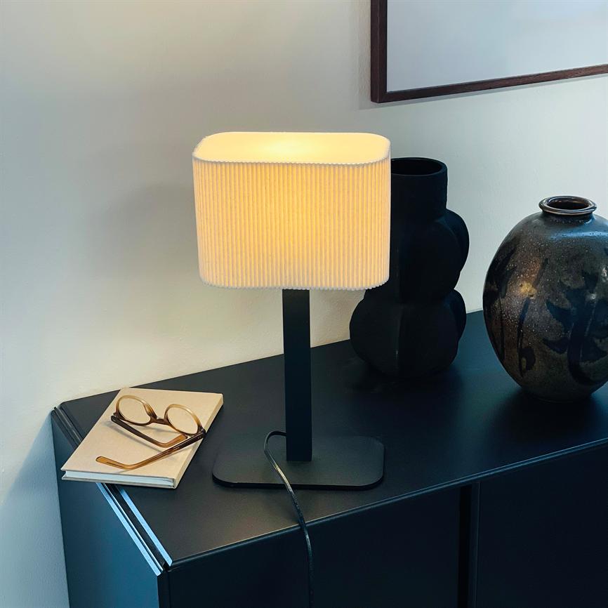 Table lamp glows softly, standing on a dark cabinet next to two decorative vases and a pair of glasses on a notebook, in front of a white wall with a framed picture.