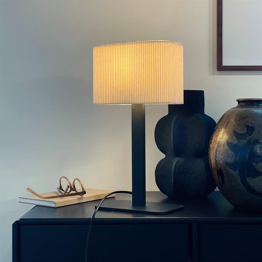 Table lamp shines warmly above a black cabinet; next to it is a stack of books with glasses, a black sculptural vase and a brown, glazed jar against a light wall.