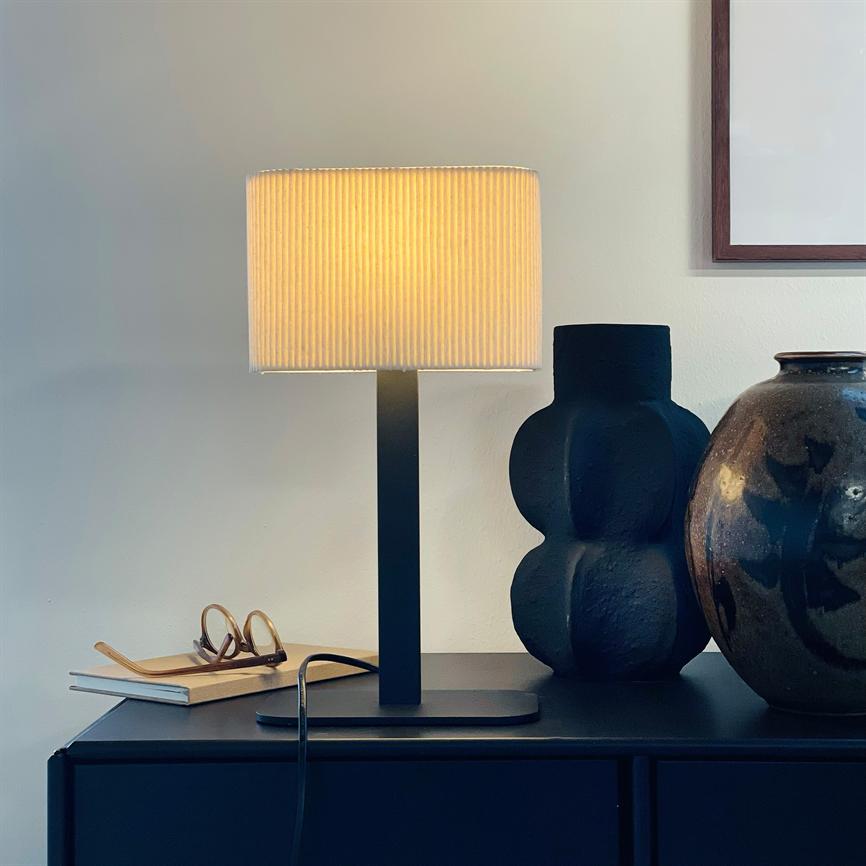 Table lamp emits warm, golden light; it sits on a dark cabinet along with a black vase, a glazed jar and a pair of glasses on a notebook beneath a framed poster.