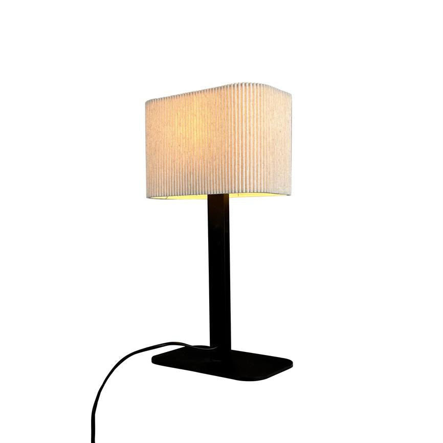 Table lamp emits warm, subdued light; stands on black rectangular base with narrow black column and light, ribbed fabric shade against white background.