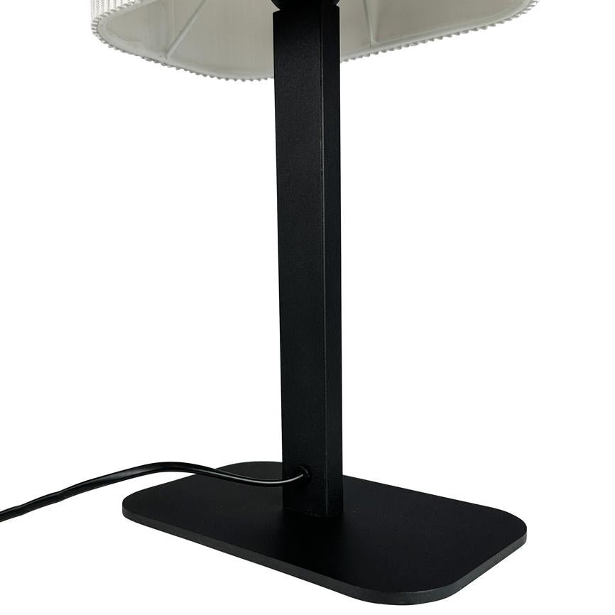 Black lamp frame stands vertically on a rounded rectangular base; a black cord runs through an opening in the foot, under a white lampshade with a wavy edge.