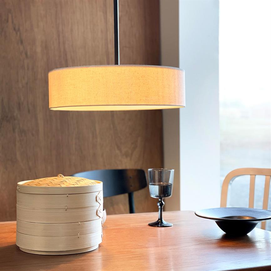 Suspended round lamp shines above a wooden table; on the table is a stack of bamboo steamers, a dark wine glass and a black bowl, with a wooden background and window to the right.