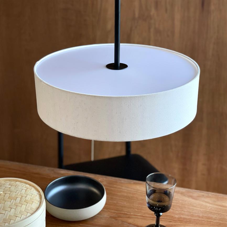 A white round lampshade hangs from a black pole above a wooden table; below it stands a white-black ceramic bowl, a bamboo lid and a dark wine glass, against a wooden wall.