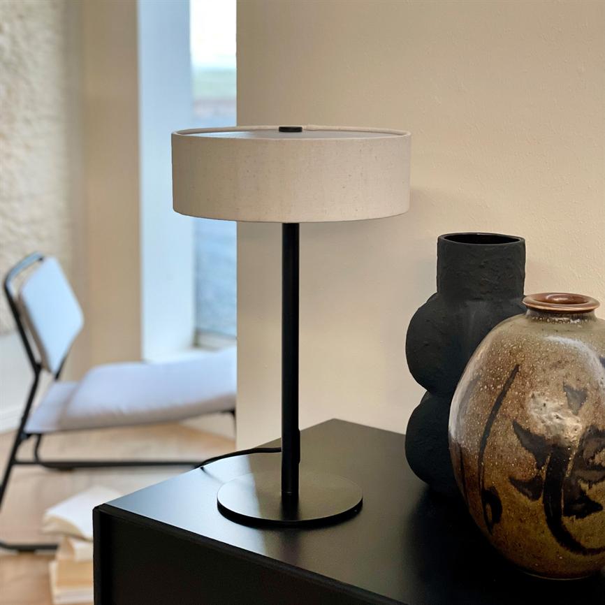 A table lamp with a black metal base and white shade stands on a black cabinet next to two ceramic vases; in the background you can see a chair and window in a bright room.