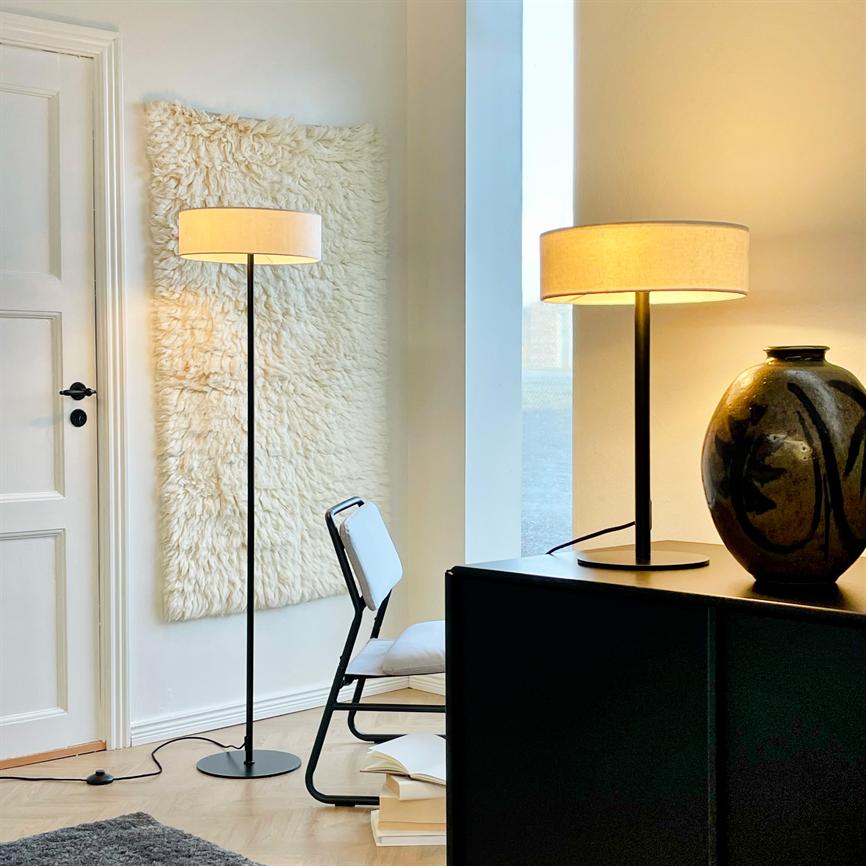 Floor lamp glows softly in a bright corner; a matching table lamp is lit on a black sideboard next to a large ceramic vase, white chair and wool rug on the wall.