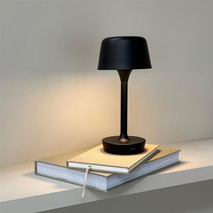Black, modern table lamp shines softly downwards and rests on two stacked books with a ribbon bookmark, placed on a simple shelf in front of a neutral, light wall.
