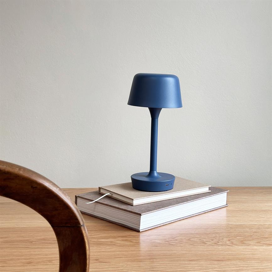Blue table lamp stands on two books on a light wooden table, with slender foot and round shade; white wall in the background and a rounded wooden chair in the left foreground.