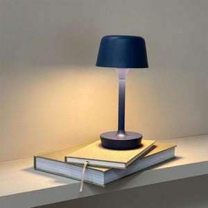 A blue table lamp is lit on two stacked notebooks; it casts warm light on a neutral wall and a simple shelf in a calm indoor environment.
