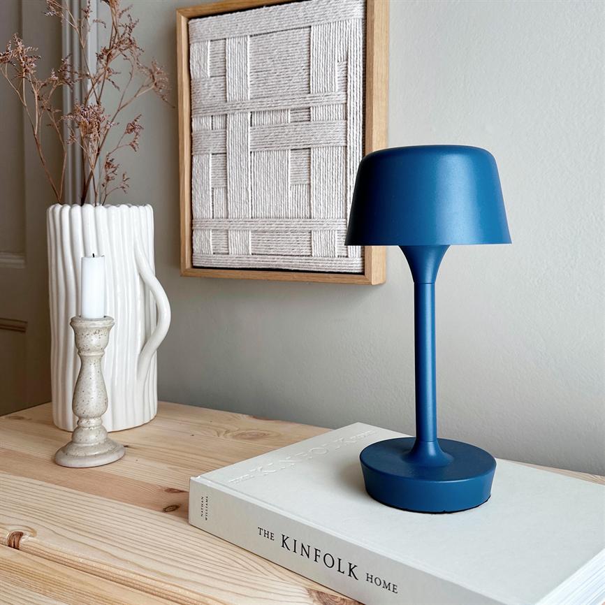 Blue table lamp stands on a book, surrounded by a light wooden chest of drawers with white vase, candle holder and framed woven art on a neutral wall. Text: "THE KINFOLK HOME".