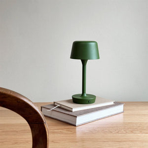 A green table lamp is switched off on a stack of books on a light wooden table in front of a light gray wall; a curved wooden chair part is seen in the foreground.
