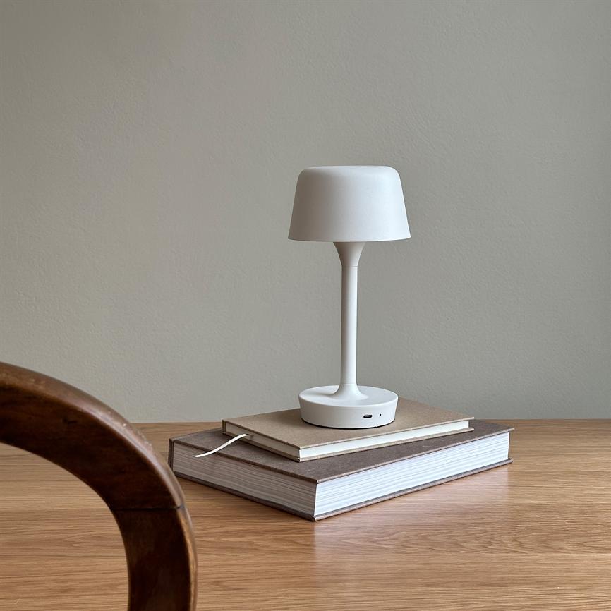 A white table lamp stands on two stacked books on a light wooden table, in front of a neutral light gray wall; in the foreground is the edge of a curved wooden chair.