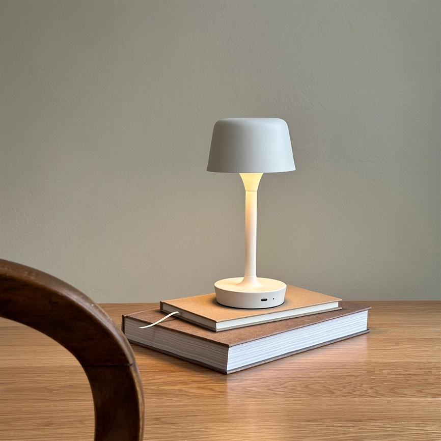 White table lamp glows softly; standing on a stack of books on a wooden table in a quiet room with light walls. A round wooden edge of a chair is seen in the foreground.