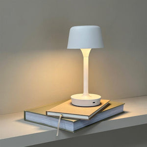 White table lamp casts soft, warm light; it stands on two stacked books on a shelf in front of a neutral, light wall.