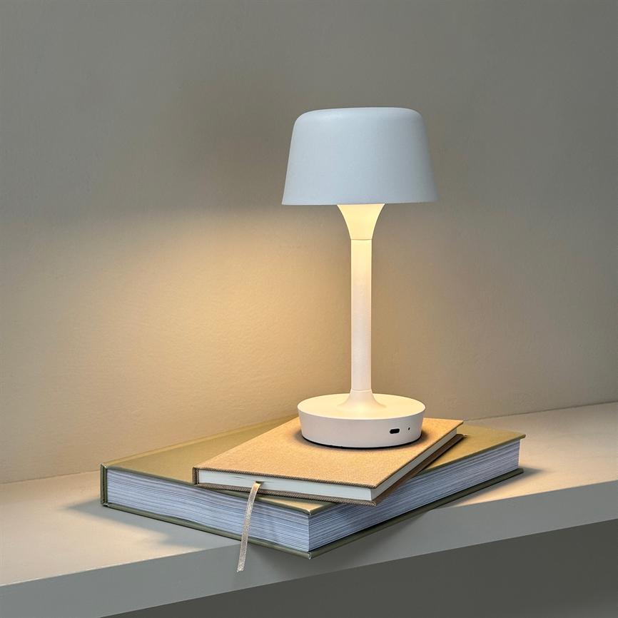 White table lamp casts soft, warm light; it stands on two stacked books on a shelf in front of a neutral, light wall.