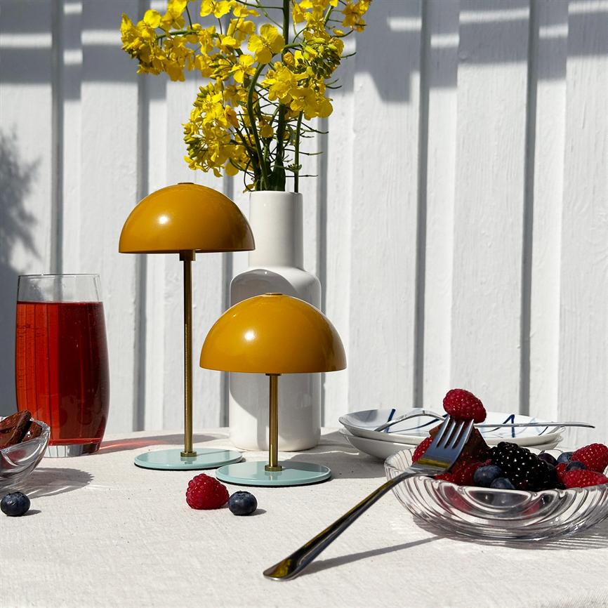 Two yellow dome lamps stand on a white tablecloth, in front of a white vase with yellow flowers; berries in a glass bowl, a fork and a glass of red drink lie on a sunlit outdoor table.