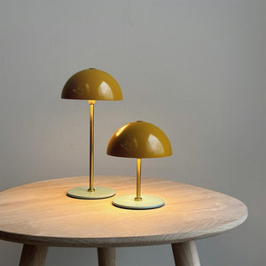 Two yellow dome lamps are lit and emit warm, soft light on a round light wooden table in a simple, neutral room with a dull gray wall.