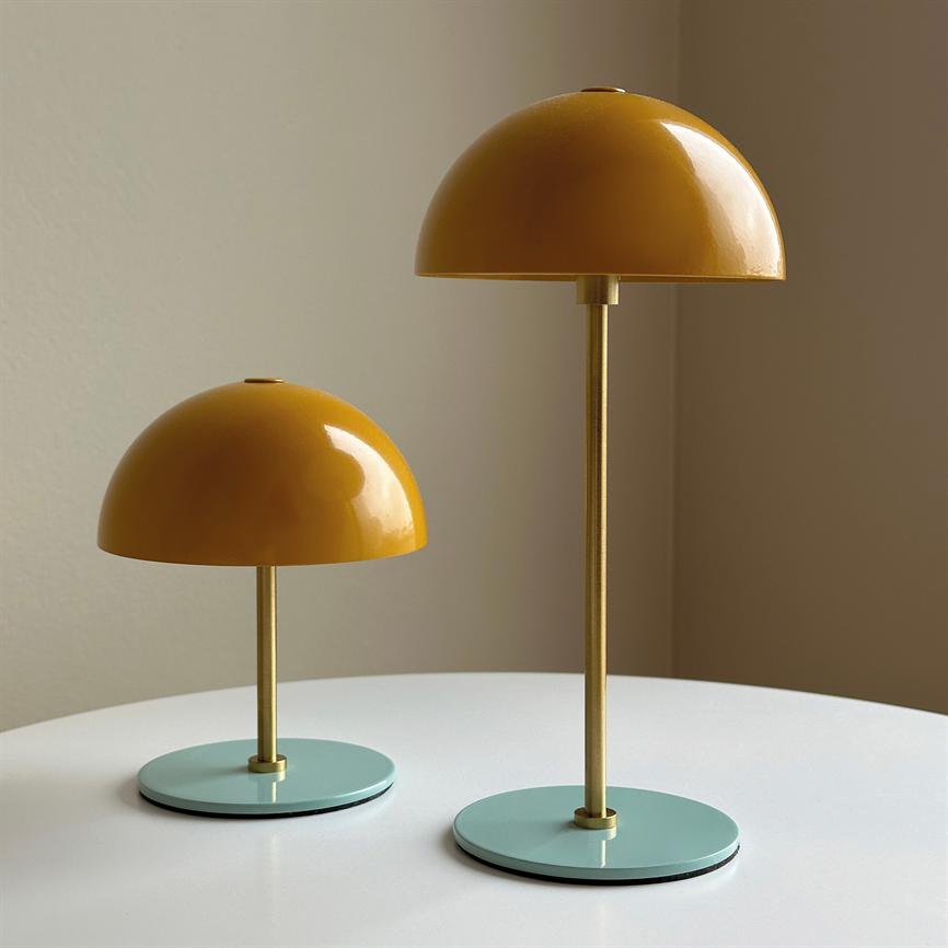Two yellow, dome-shaped lamps stand on a round white table; one taller than the other, with golden stems and mint green round plinths in front of a beige wall.
