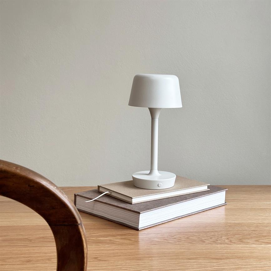A white table lamp rests on two stacked books on a light wooden kitchen table, with a curved wooden chair in the foreground and a neutral beige wall in the background.