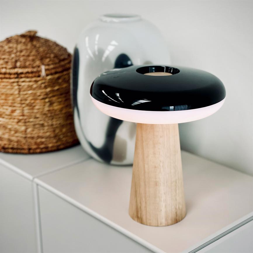 Table lamp with wooden base and round, black glossy shade emits soft pink light; stands on a white dresser in front of a shiny silver vase and wicker basket in a bright, minimalist room.