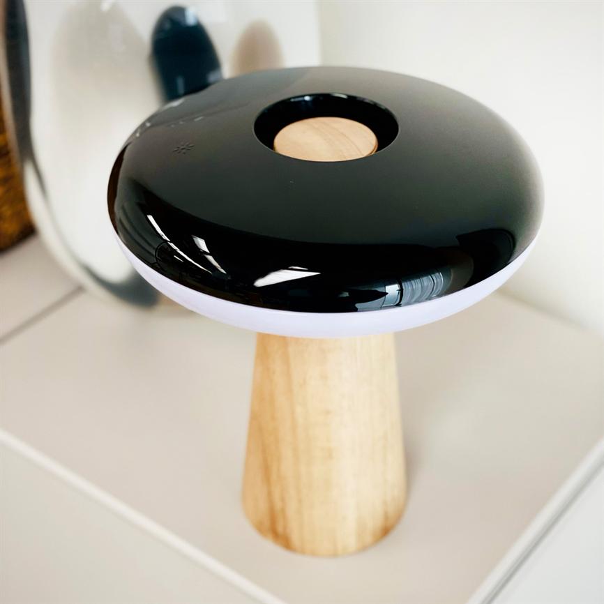 A black, glossy, mushroom-like lamp with a round wooden insert and white rim stands on a light wooden base on a white table top and is reflected in a mirror behind it.