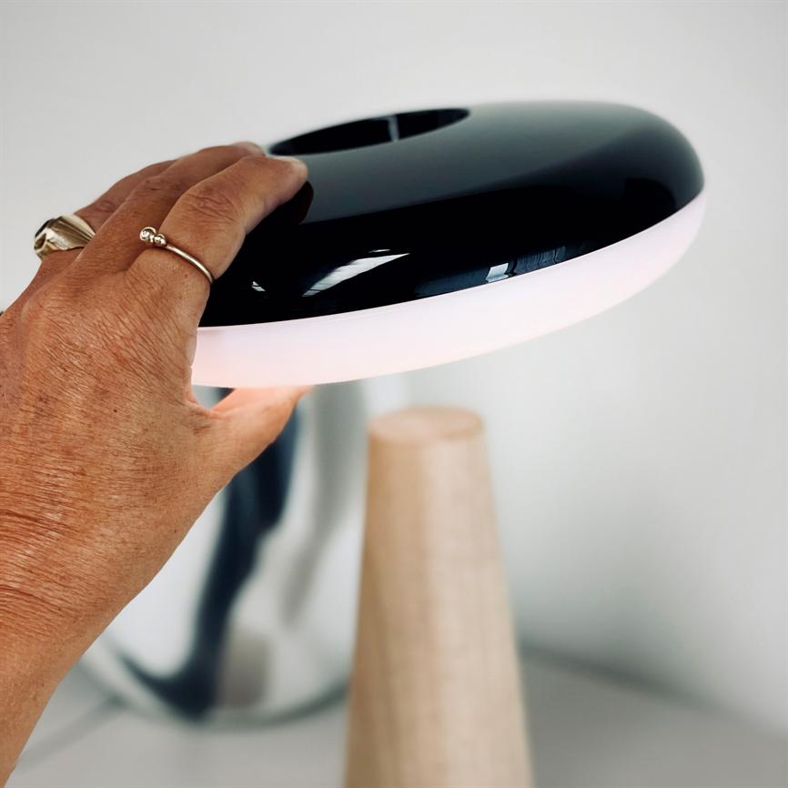Table lamp — a hand lifts the black, round shade with a luminous white border; the lamp stands on a cone-shaped wooden base in a bright, minimalist room.