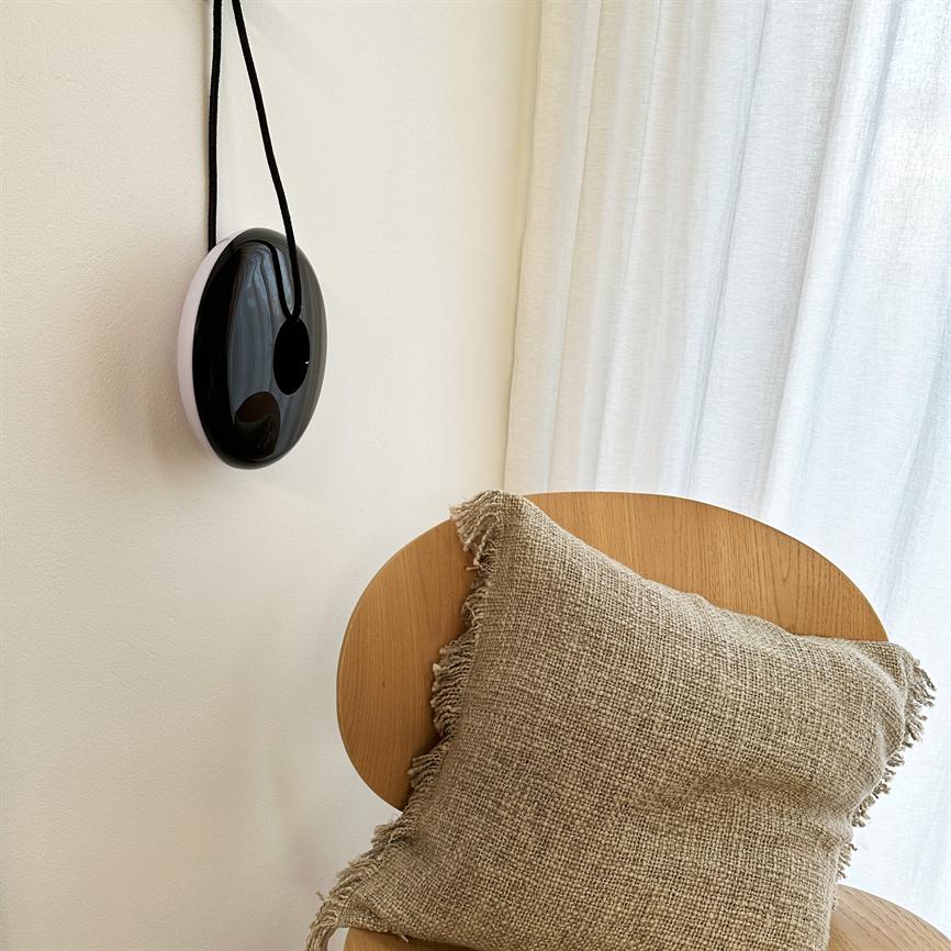 Black oval object hangs on a black string on a light wall, next to a wooden chair with a coarse beige decorative pillow and light curtains in the background.