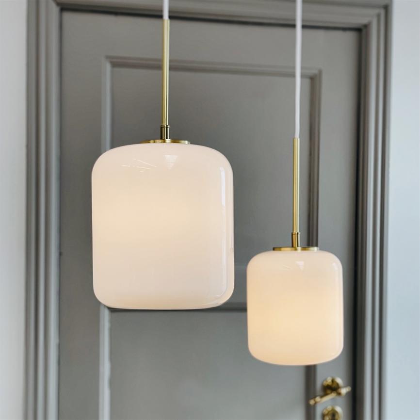 Two lighted, milky-white pendant lamps in a rounded square shape hang from brass shafts, in front of a gray infill door in a bright room; a brass doorknob is seen in the lower right.