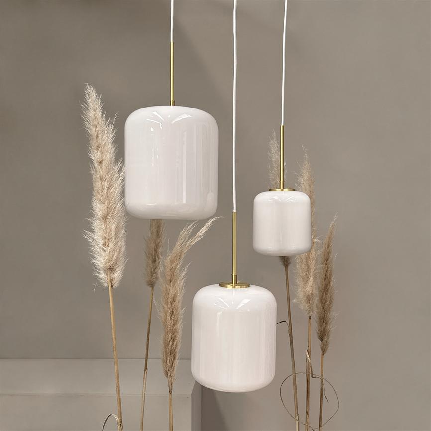 Three white cylindrical pendant lamps with brass fittings hang at different heights; bordered by dry pampas grass stalks in front of a neutral sand colored wall.