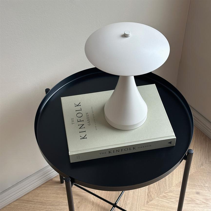 A small white table lamp stands on a closed book on a round black side table in a bright corner by the wooden floor and wall. Text on the book: "THE KINFOLK" "GARDEN" (spine: "THE KINFOLK GARDEN").