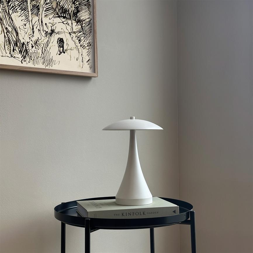 A white, mushroom-like table lamp stands on a book on a round, black side table in a bright, minimalist corner. Text on spine: "THE KINFOLK GARDEN".