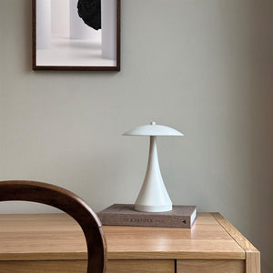 White table lamp stands on a book on top of a light wooden desk; round wooden chair in the foreground, neutral wall and framed art hanging above. Text on book: THE KINFOLK TABLE.