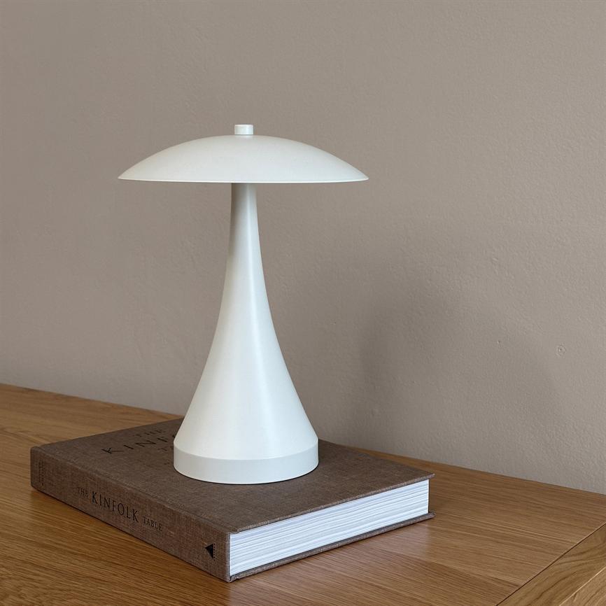 White, mushroom-shaped table lamp stands on a thick, brown book placed on a light wooden table in front of a plain beige wall. 
 Text on spine: THE KINFOLK TABLE.