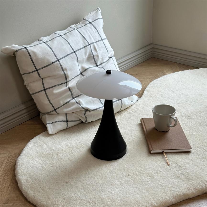 A black, mushroom-like table lamp stands on a soft, white fur rug; next to it is a checkered pillow, a brown notebook and a gray ceramic cup in a quiet corner.