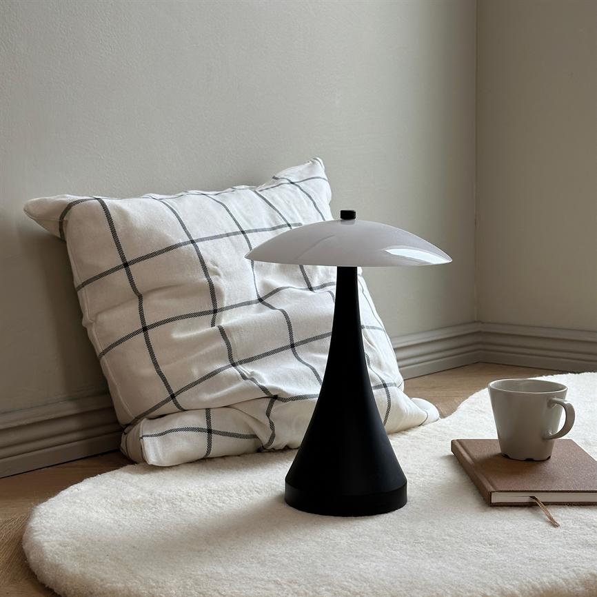 A black, cone-shaped table lamp with a white dome stands in the middle of a soft round white rug; a checkered pillow leans against the wall, next to it is a notebook and a coffee cup.