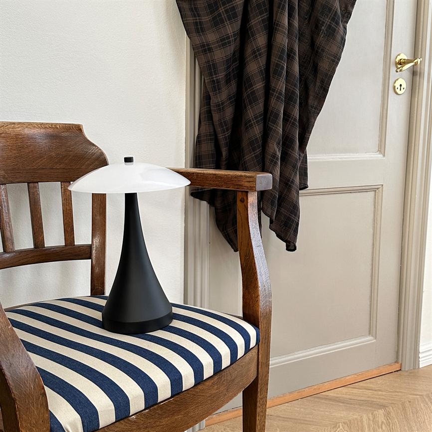 Black conical table lamp with white shade stands on a wooden chair. A blue-and-white striped seat, a checkered jacket hanging on the door and a light wooden floor can be seen around.