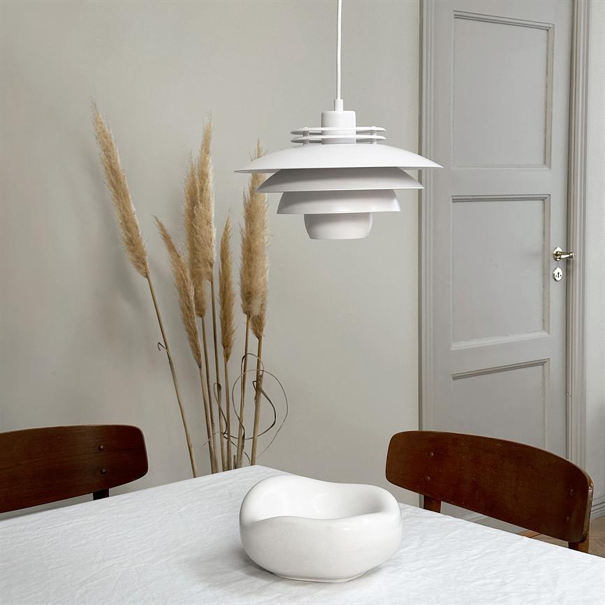 A white, layered pendant hovering over a white-clothed table; an organic white bowl sits on the table, surrounded by two wooden chairs, dried pampas grass and a closed door.