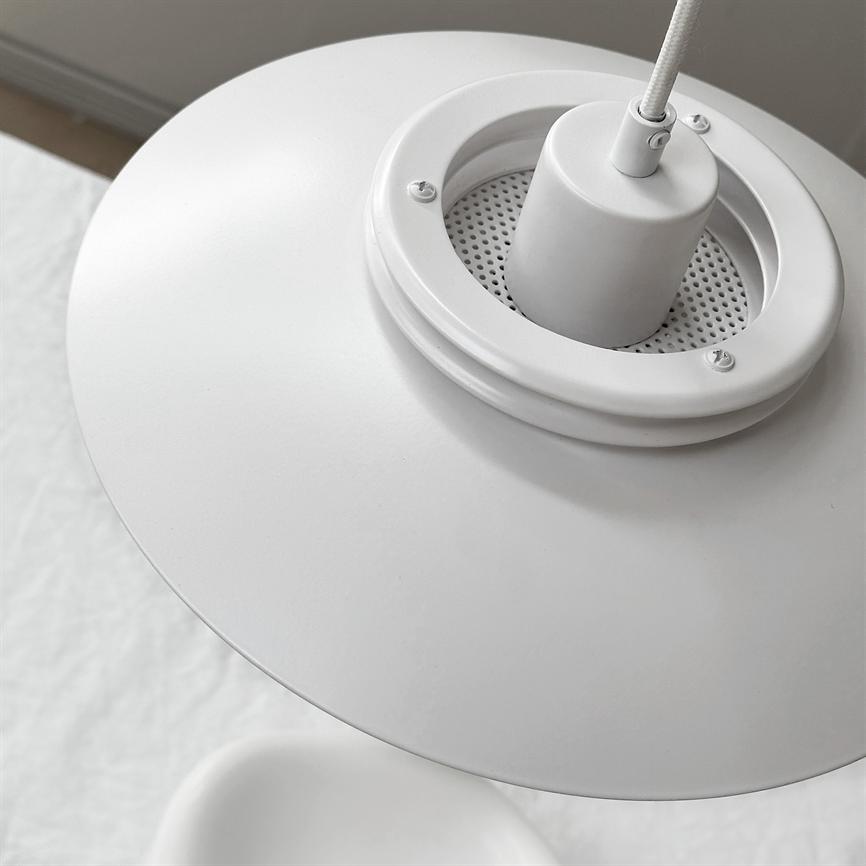 A white metal pendant lamp hangs from a fabric-covered cord; the lamp's round shade and perforated socket are seen obliquely. Context: bright, minimalist interior with white textile and neutral wall.