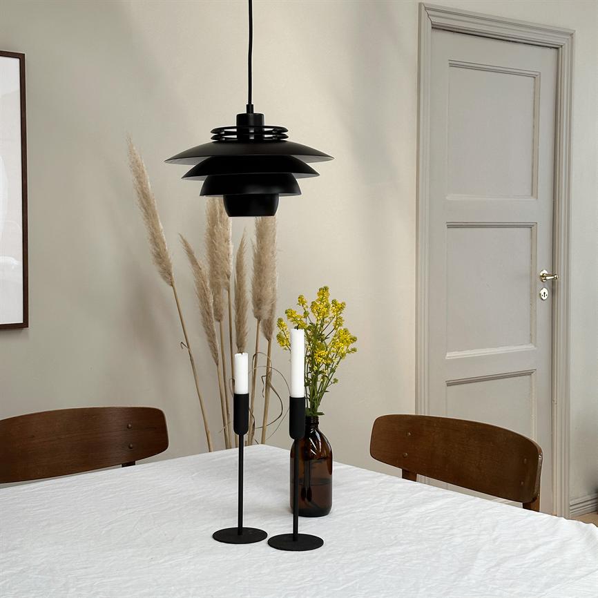 A black pendant lamp hangs above a white, dew-covered table with two tall black candlesticks and a brown bottle with yellow flowers, surrounded by brown chairs, pampas grass and a closed door.