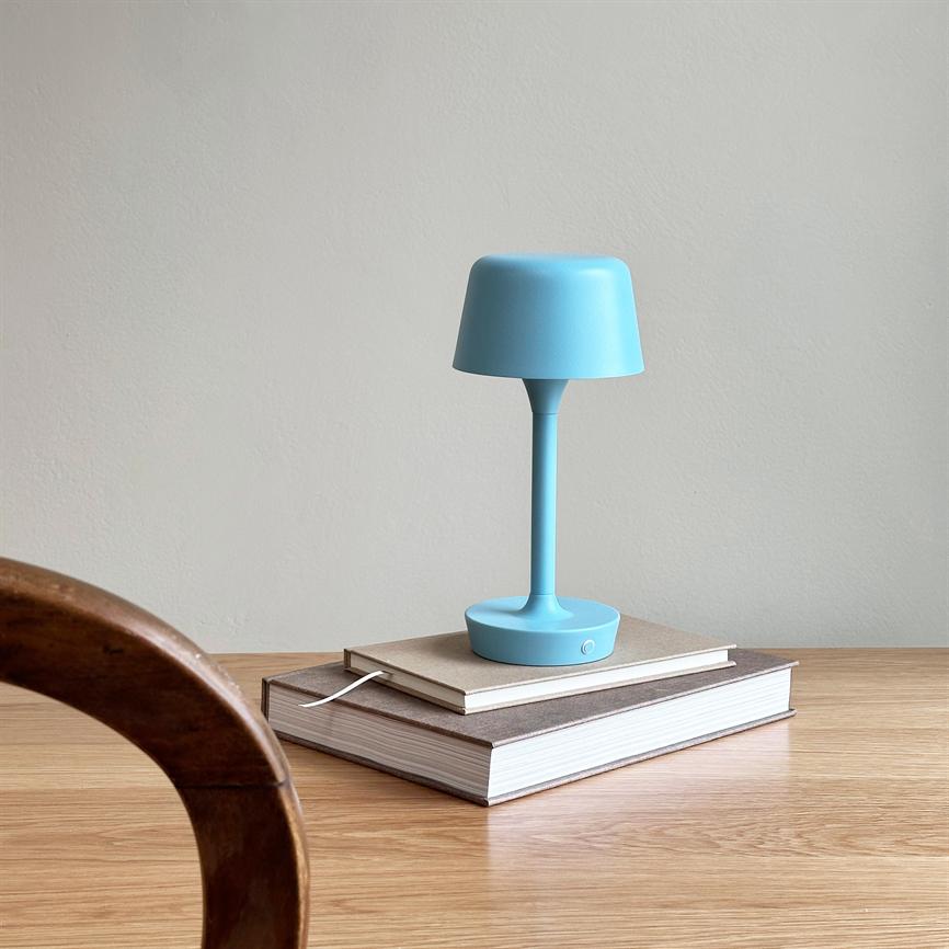 A blue table lamp sits on a stack of books on a light wooden table in front of a light gray wall; a curved chair back is seen in the foreground.