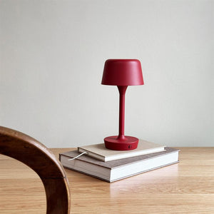 Red table lamp stands on two stacked books on a light wooden table; a brown wooden chair is seen in the foreground, simple white wall in the background.