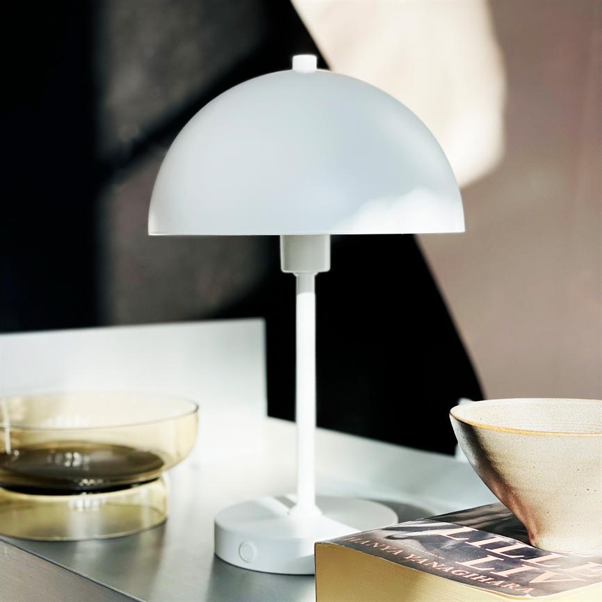 A white, dome-shaped table lamp stands on a table; next to two transparent glass bowls, a ceramic bowl and a book on a light surface in front of a dark background.