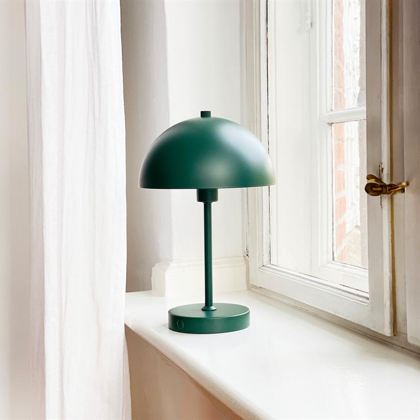 Green metal table lamp stands on a white windowsill by a window; soft daylight filters in, in front of a light curtain and an open casement window.