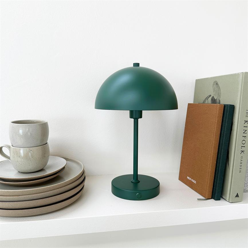 A dark green table lamp is switched off on a white shelf, flanked by beige plates and cups on the left and books on the right. Text: "THE KINFOLK GARDEN"; "NEW MAGS".