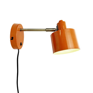 An orange wall lamp mounted on a turned metal arm, shines downwards; round wall plate and black wire visible against a neutral white background.