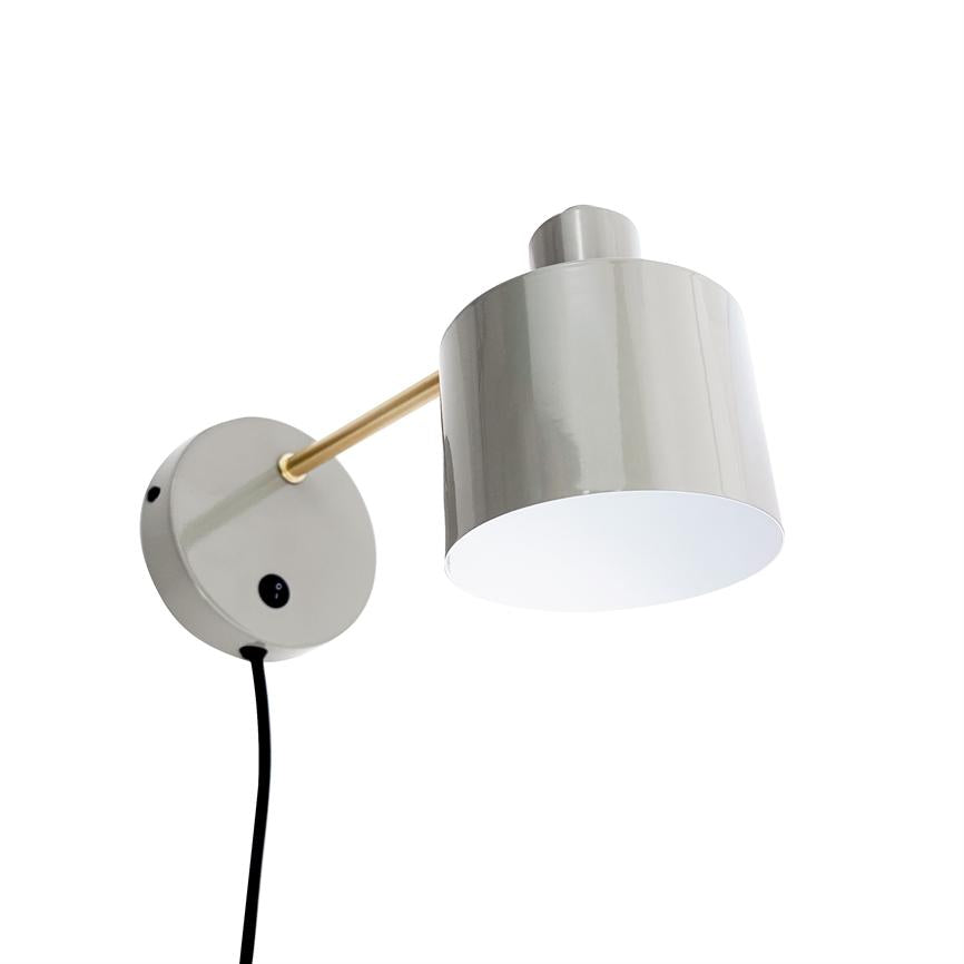 Gray cylindrical wall lamp with gold colored arm mounted on round wall bracket, a black cord hangs down; the lampshade points downwards against a neutral white background.