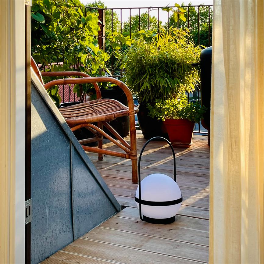 A round, white portable lantern stands on the wooden floor of a small balcony; surrounded by a rattan armchair, green potted plants, a metal railing and soft evening light.