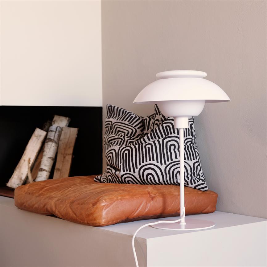A white table lamp stands on a white shelf; next to it is a brown leather cushion and a black and white patterned cushion, in front of a fireplace opening with stacked birch logs.