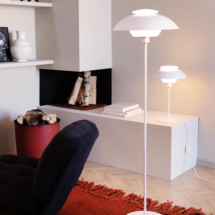 A white floor lamp glows softly; a smaller matching table lamp sits on a low shelf. Surroundings: built-in fireplace with firewood, red firewood basket, dark armchair and red carpet.