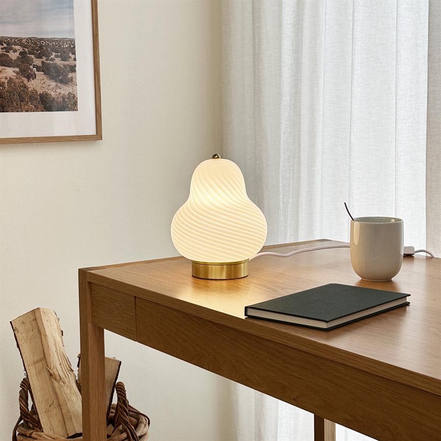 Table lamp with white, wavy glass is lit on a wooden table and emits warm light; next to it is a closed notebook and a mug in front of bright curtains and wall art.